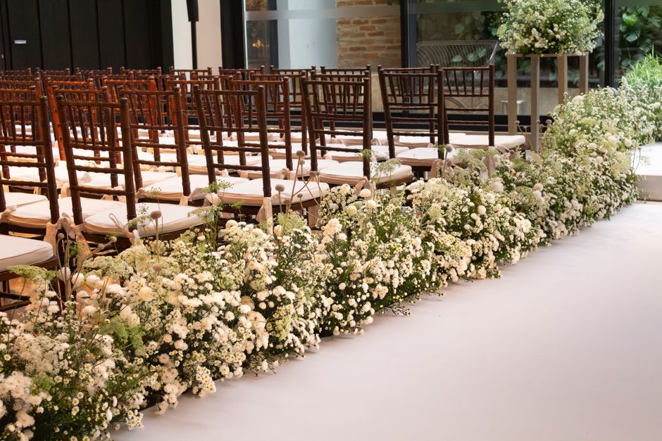 A row of wedding chairs with white cushions, arranged in a grand indoor venue, each positioned behind a lush floral arrangement consisting of small white roses, baby's breath, and green foliage. The f