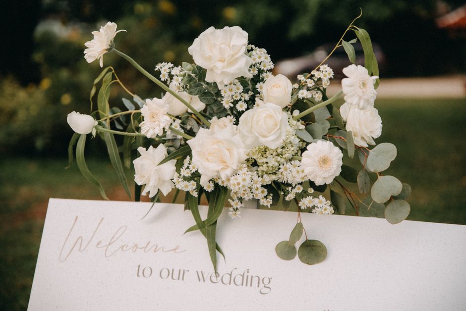 A bouquet arrangement of predominantly white flowers, including large roses, delicate daisies, and small filler blooms, accented with eucalyptus leaves and long, slender greenery. The flowers are fres